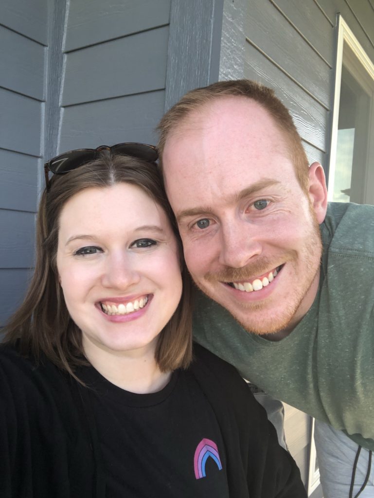 A couple posed with their house in the background - all you can see is siding. Both are white, and the woman has a bi pride shirt on; she's being embraced by her husband. 