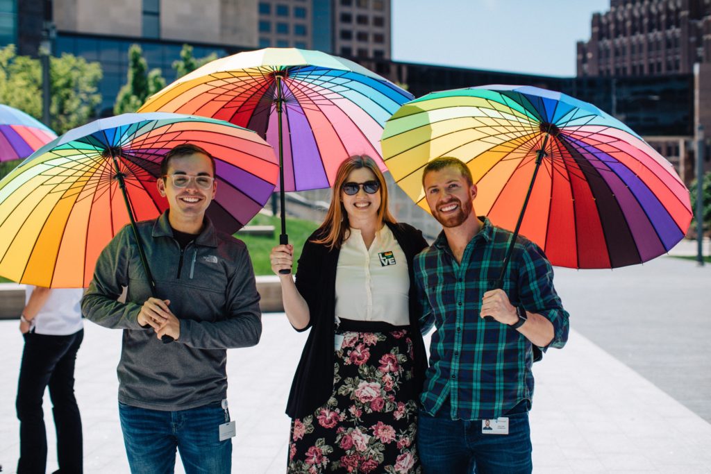 Three people holding large rainbow umbrellas outdoors. 